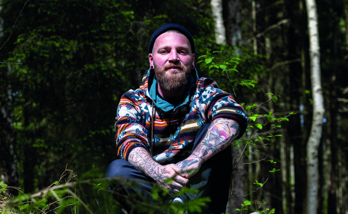 Ein Mann mit Tattoos und bunter Kapuzenjacke sitzt im Wald, bereit für eine Achtsamkeitssitzung mit einem Naturcoach.