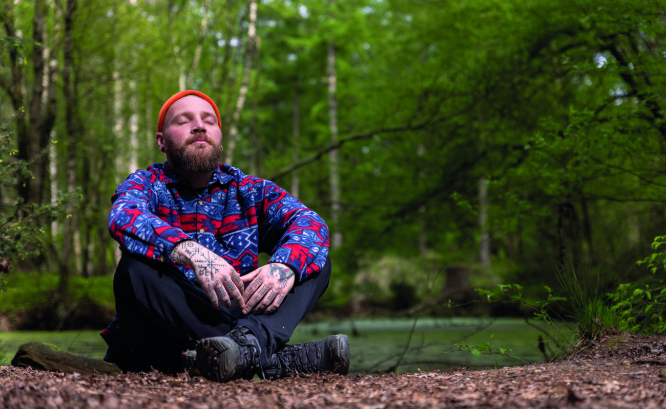 Ein bärtiger Mann mit Tattoos, der einen blau-roten gemusterten Kapuzenpullover und eine orangefarbene Mütze trägt, sitzt im Schneidersitz in einem ruhigen Waldgebiet. Er hat die Augen geschlossen und scheint zu meditieren. Der Hintergrund ist mit grünen Bäumen und einem kleinen Teich gefüllt, was eine friedliche natürliche Umgebung schafft.